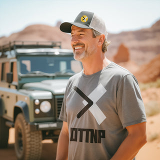 A man standing in the Utah desert next to his old Land Rover Defender wearing a black and white FlexFit hat with our OTTNP circle logo. The man is also wearing our grey black and white OTTNP logo t-shirt. ottnp.com